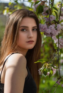 Siyah elbiseli havalı esmer kız, Sakura ağacının yanında kameraya bakarak poz veriyor. Dikey. 