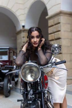 Uzun siyah saçlı seksi kız tarihi bir motosikletle poz veriyor. Dikey