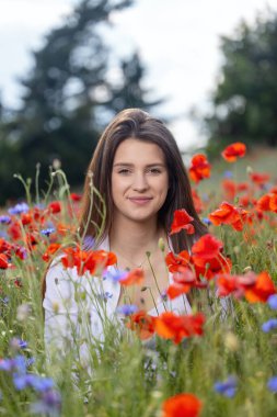 Gülümseyen uzun saçlı esmer kız, çayırda kırmızı gelincikler ve mavi çiçeklerle poz veriyor. Dikey. 