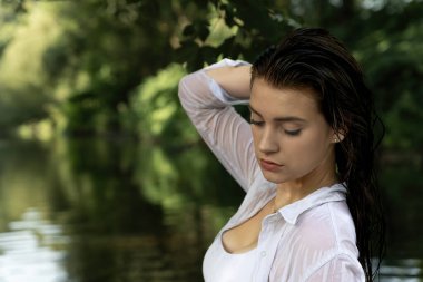 Nehirde ıslak saçlarla poz veren baştan çıkarıcı genç bir kızın portresi. Kız tek eliyle saçına dokunuyor. Yatay olarak. 