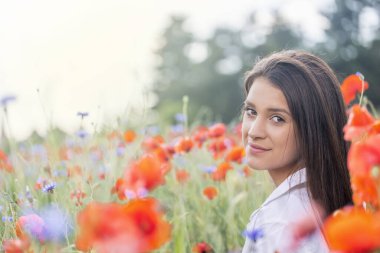 Güzel esmer, genç bir kadın kameraman kırmızı gelincik tarlasında omzunun üzerinden bakıyor..