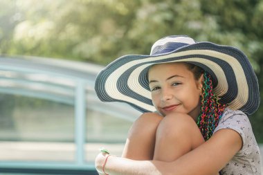 Hasır şapkalı havalı küçük kız havuzun önünde oturuyor. Yatay olarak. 