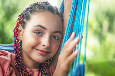 Saçında renkli örgüler olan genç ve güzel bir kız mavi hamakta poz veriyor. Yatay olarak. 
