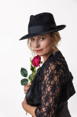 Siyah dantelli bir elbise ve elinde kırmızı bir gül tutan orta yaşlı bir kadın beyaz bir arka planda poz veriyor..