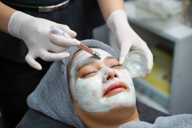 Kapalı kozmetik uzmanı güzellik kliniğindeki kadın hasta yüzüne yüz maskesi kremi sürüyor..