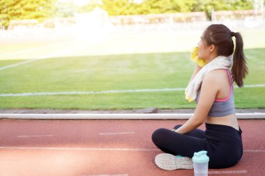 Young Asian female relaxing after workout and jogging at the football stadium in the morning with sun flare background.