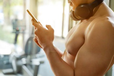 Yaklaş ve doğra Asyalı adamın mükemmel vücudu. Parlak güneş ışığı arka planında fitness merkezinde cep telefonuyla rahatlıyor..