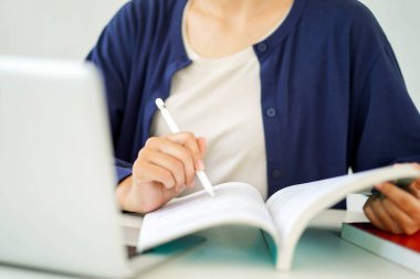 Yakın plan ve günlük giysiler içinde genç bir kız öğrenci bulanık arkaplan sınavı için dizüstü bilgisayarla kitap okuyor..