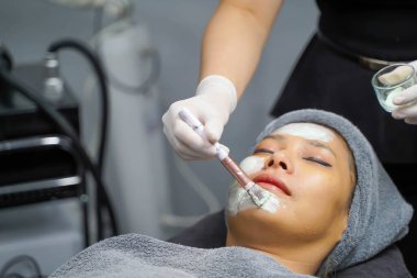 Kapalı kozmetik uzmanı güzellik kliniğinde kadın hasta yüzüne yüz maskesi kremi sürerken kozmetik fırçası kullanıyor..