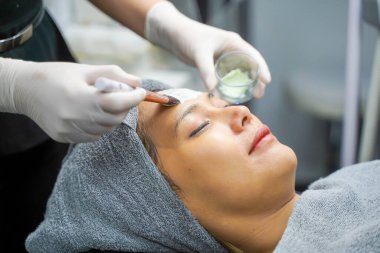 Kapalı kozmetik uzmanı güzellik kliniğinde kadın hasta yüzüne yüz maskesi kremi sürerken kozmetik fırçası kullanıyor..