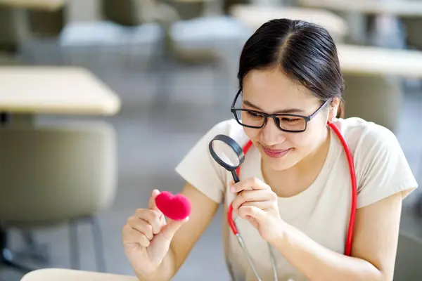 Medical student holding a magnifying glass and looks at a yarn red heart on blurred background. Asian medical student with Heart disease concept.