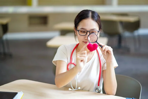 Medical student holding a magnifying glass and looks at a yarn red heart on blurred background. Asian medical student with Heart disease concept.