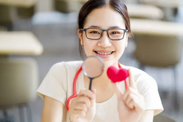 Closeup medical student action holding a magnifying glass and a yarn red heart with cute smile on blurred background. Asian medical student with Heart disease concept.