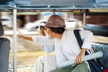 Yakın plan Amerikalı turist Tuktuk Tayland taksisine biner ve tur rehberiyle şehirdeki ilginç yerleri gösteren bir fotoğraf çeker. Kıdemli turist konsepti