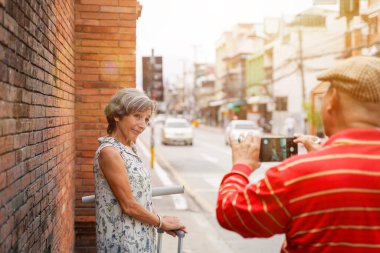 Yakın plan kıdemli bayan turist, arkadaşının şehirde güneş ışığında yürürken fotoğraf çekmesi ve şehrin arka planının bulanıklaşması için poz veriyor..