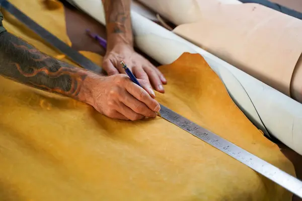 Closeup and crop hands of leather craftsman using a pencil and steel ruler drawing design on the yellow leather in the workshop.