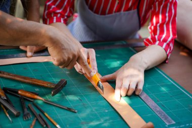 Kıdemli zanaatkârın genç kadın deri imalatçısına atölyesinde kemer yapmak için deri kesme bıçağı kullanmayı öğrettiği yakın plan ve mahsul eli..