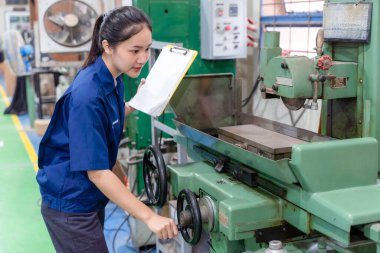 Üniforma giyen Asyalı endüstriyel mühendis bir pano tutuyor ve makinenin endüstriyel atölyedeki eski model torna ve matkap işletim sistemini kontrol ediyor..