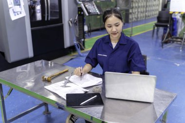 Endüstriyel atölyede dizüstü bilgisayarla makine parçaları mühendisliği yapan Asyalı kadın teknisyen..