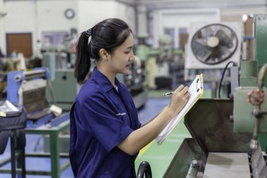 Asyalı endüstriyel mühendis kadın bir panoya bakıyor ve endüstriyel atölyedeki eski model torna ve matkap makinesini kontrol ediyor..