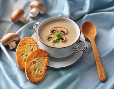 Biraz mantarlı kremalı mantar çorbası, sarımsaklı ekmekle süslenmiş bahar soğanı ve ahşap kaşık zarif bir şekilde mavi ipek arka planda mavi ipekli beyaz seramik bardakta servis edilir.