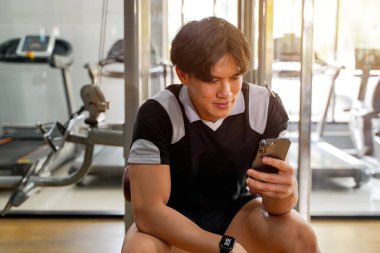Yakın plan Asyalı yakışıklı çocuk spor yaparken cep telefonuyla güneşli arka planda spor salonunda dinleniyor..