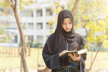 Yakından Müslüman bir öğrenci, abaya niqab giyiniyor geleneksel giysiler giyiyor ve okul binasında güneş ışığıyla parlayan Kuran okuyor..