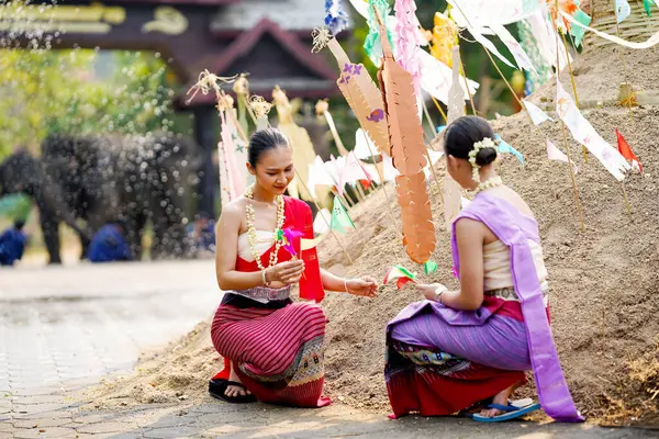 Tayland geleneksel elbiseli güzel Taylandlı kadınlar Songkran festivalinde tapınaktaki kum yığınına üçgen bayrak dikmeye yardım ediyorlar..