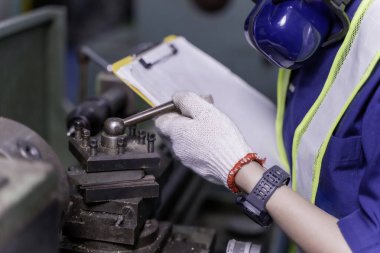 Eski model torna ve matkap makinelerini kontrol eden ve endüstriyel atölyedeki panoya bir bakım listesi yazan, üniformalı endüstriyel mühendisin yakın çekim ve tepeleme eli..