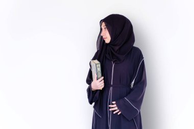 Güzel bir Müslüman öğrenci abaya niqab geleneksel kıyafet portresi giyiyor ve beyaz duvar arkasında Kur 'an' ı tutuyor. Arapça metinler İngilizce 'de şanlı Kuran' dır..