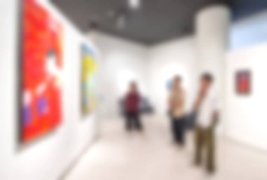 Sanat sergisi arka planı bulanık ve insanlar beyaz duvardaki resimlere bakıyor.