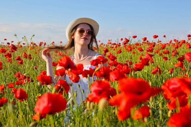 Haşhaş tarlasında şapkalı ve güneş gözlüklü güzel bir genç kızın portresi. Doğada güzellik, kırsal alan, genç kız ve çayırda bir sürü kırmızı çiçek, altın saatinde ilkbahar günü