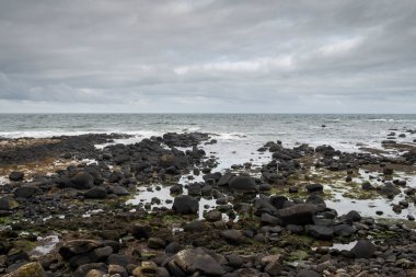 Kuzey İrlanda 'daki Giant' s Causeway yakınlarında Antrim Sahili boyunca uzanan siyah bazalt çakıl taşı plajından sakin bir manzara. Ufuk bulutlu bir gökyüzünün altında okyanus boyunca uzanıyor.