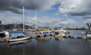 Kuzey İrlanda, Belfast 'taki Lagan Nehri' nde demirlemiş tekne ve yatlar, modern binalar ve arka planda parçalı bulutlu bir gökyüzü.