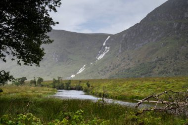 A scenic view of Lough Veagh, in Ireland, with the Astelleen Burn Waterfall cascading down a mountain slope, surrounded by lush greenery and reflecting the overcast sky, creating a serene and natural atmosphere clipart