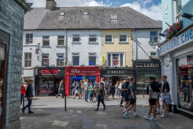 GALway, IRELAND - 6 AĞUSTOS 2023: Galway 'de hareketli bir ticari cadde, dükkanlar boyunca yürüyen insanlar, canlı mağazalar ve parçalı bulutlu bir gökyüzü altında canlı atmosfer