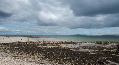 Silverstrand Sahili 'nde çakıl taşlı sahil, Galway, İrlanda, sakin deniz, parçalı bulutlu gökyüzü ve kumsalın keyfini çıkaran insanlar.