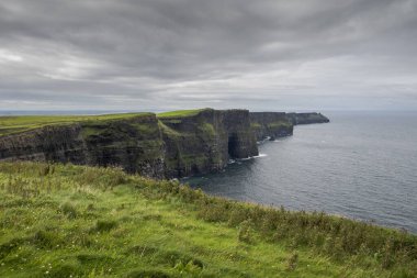 İrlanda 'nın batı kıyısındaki Moher Kayalıkları' nda gri gökyüzünün altındaki uçurumun kenarına uzanan yeşil çimenler doğal manzarayı ve jeolojik özellikleri gözler önüne seriyor.