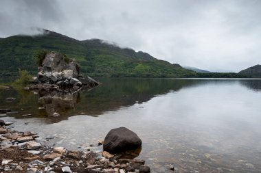 İrlanda 'nın Muckross Gölü' nün sakin bir sahnesinde sergilenen sergiler bulutlu gökyüzünü yansıtıyor, arka planda kayalık bir kıyı ve sisli tepeler, sakin ve resimsel bir manzara yaratıyor.