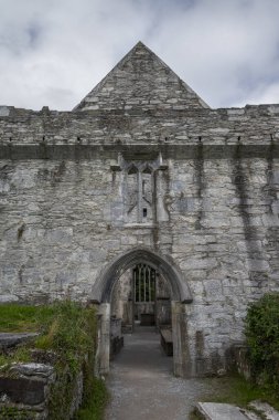 İrlanda 'daki Muckross Manastırı cephesinde yıpranmış taş duvarlar ve bulutlu gökyüzünün altında sivri bir kemer girişi yer alıyor.