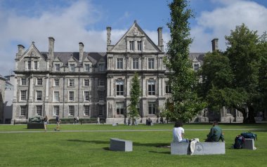 DUBLIN, IRELAND - 10 AĞUSTOS 2023: Dublin 'deki Trinity Koleji kampüsünde insanların parçalı bulutlu bir gökyüzü altında oturup yürüdükleri yeşil çimenlikli bir bina manzarası.