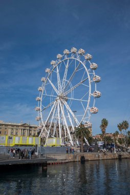 BARCELONA, İspanya - 7 Aralık 2023 Barcelona 'nın eski limanının önünde büyük bir dönme dolap. Beyaz yapı mavi gökyüzü ile çelişiyor, ve insanlar iskele boyunca yürüyor, yakınlarındaki palmiye ağaçları ve tarihi binalarla.