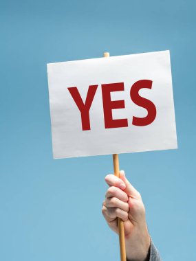 Closeup of a male hand holding a white sign with word YES in red letters. on blue background clipart