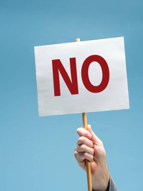 Closeup of a male hand holding a white sign with word NO in red letters. on blue background clipart