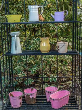 Açık-çerçeve dizaynının metal rafı çeşitli renkli sulama kutuları, sürahiler ve küçük kovalarla dolu, açık havada, yemyeşil yapraklarla çevrili, canlı bir bahçe sahnesi yaratıyor.