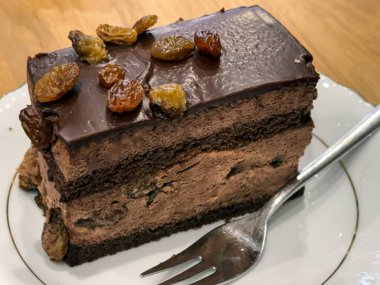 Kuru üzümle kaplı zengin çikolatalı pasta dilimi, çatalın yanında beyaz bir tabakta servis ediliyor.