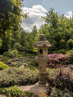 Geleneksel taştan bir fener, çiçek açan bir gölet ve parlak bir gökyüzünün altında yemyeşil bir gölet içeren güzel bir bahçe.
