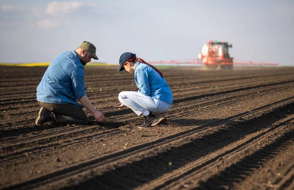 Mladí Farmáři Zkoumají Nečistoty Zatímco Traktor Rozprašuje Pole — Stock fotografie