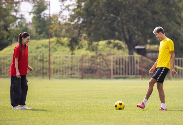 Genç bir futbolcu, genç bir kıza futbol sahasında güneşli bir günde topa nasıl vurulacağını öğretiyor.