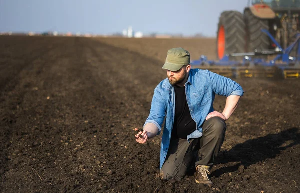 Young Farmer Examing Dirt While Tractor Plowing Field Stock Photo